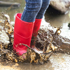A pair of red Classic Wellies are shown splashing in a puddle, covered in mud, with blue jeans worn above.